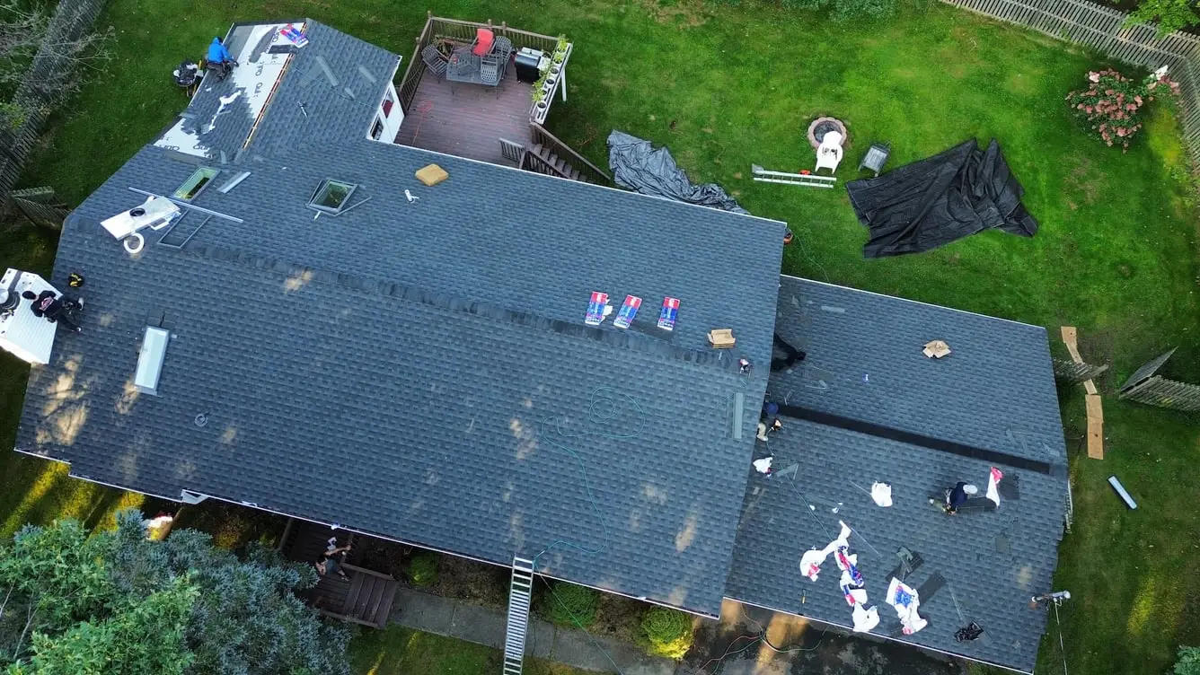 Aerial view of a newly shingled roof on an L shaped house, with tarps and debris still on the lawn